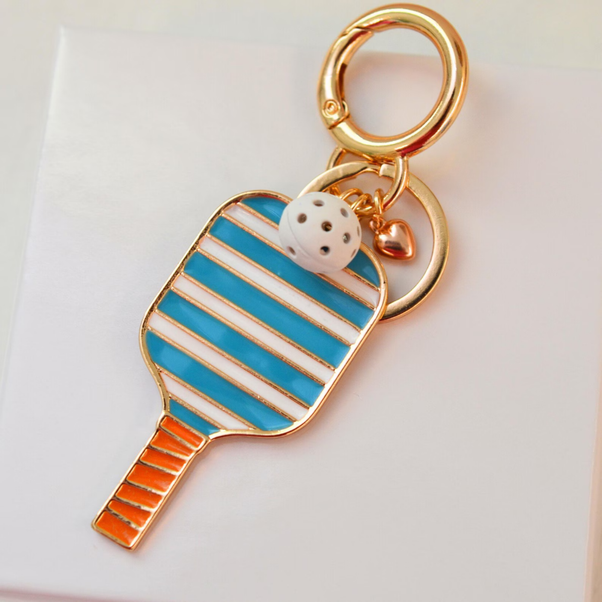 A tennis racket-shaped keychain charm with gold-plated metal trim, turquoise blue and white striped enamel, an orange grip handle, a white die-cut tennis ball charm, and a small gold heart accent, displayed on a clean white background.