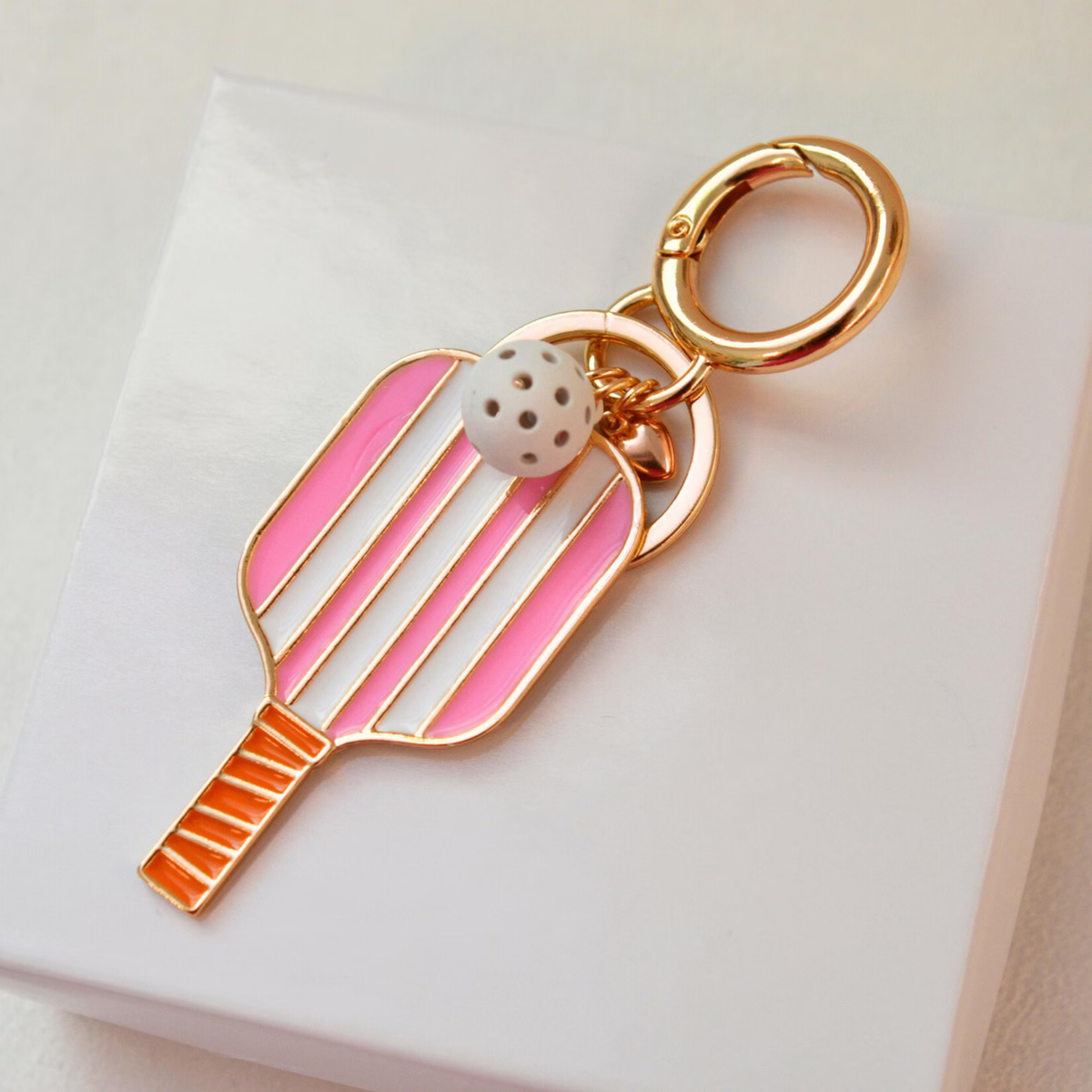 A gold-tone tennis racket keychain charm with pink and orange enamel details, featuring a small white beaded ball accent, displayed on a clean white surface.