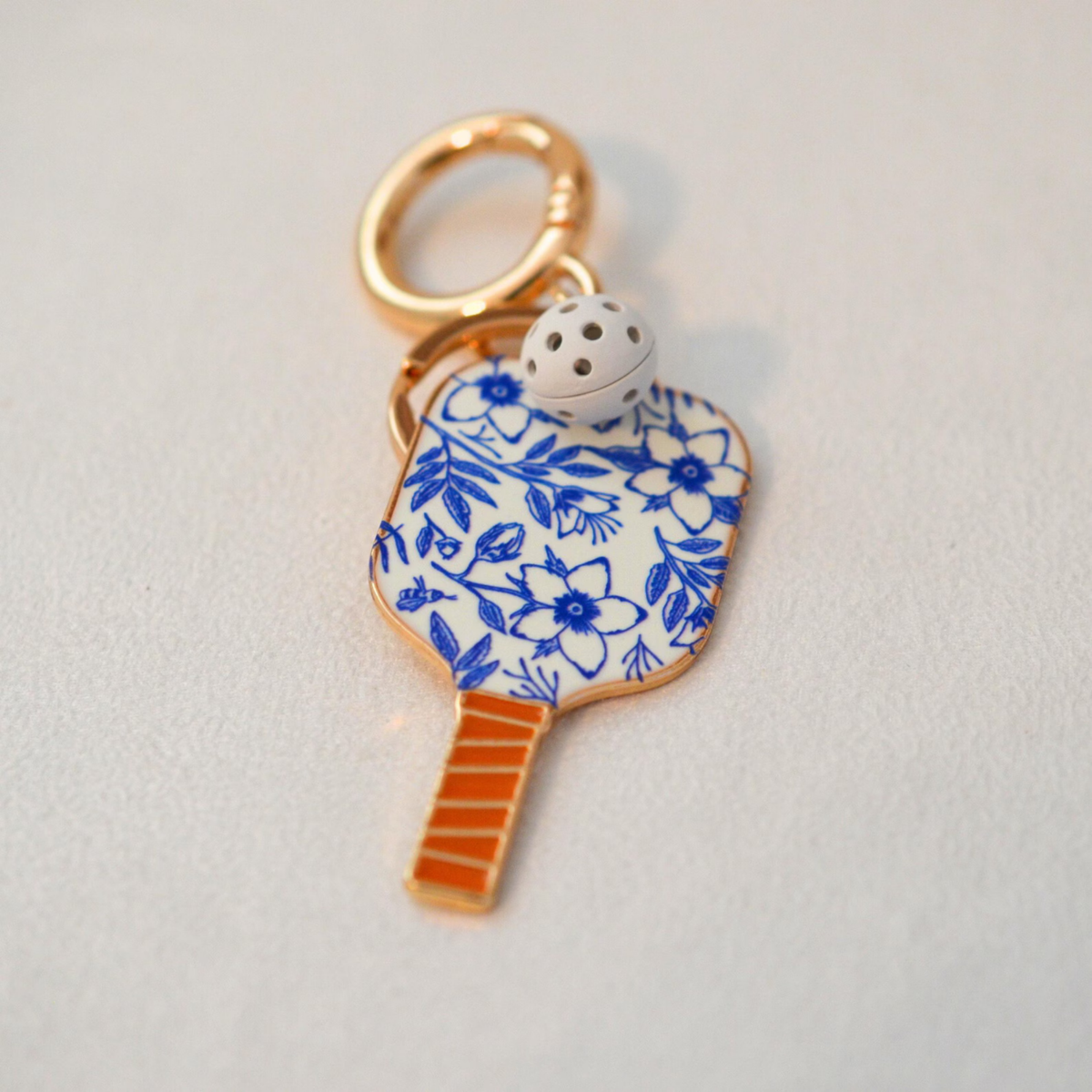 A close-up shot of a decorative keychain charm shaped like a paddle racket, featuring a blue and white floral pattern, a gold-tone handle, and a small white bell with black dots, set against a plain light gray background.