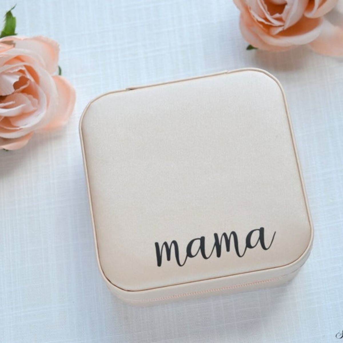 A square cream-colored jewelry box with rounded corners and the word 'mama' printed in black script on the front, styled on a white textured surface with pink roses in the corners.