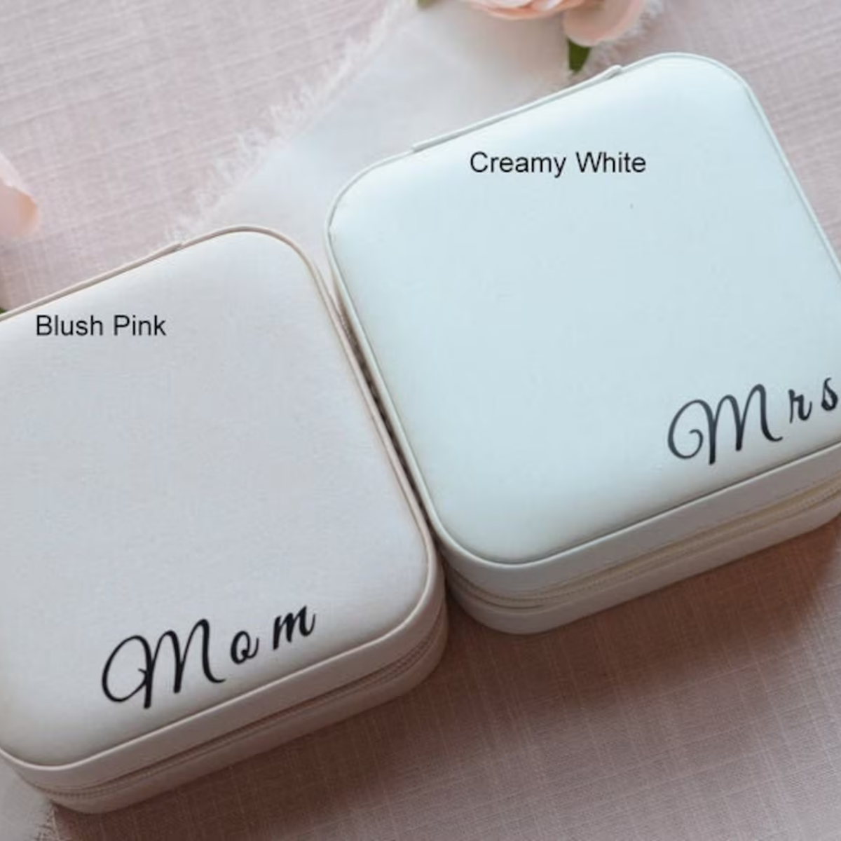 Two small square zippered jewelry boxes shown on a soft pink fabric background. One box is labeled 'Blush Pink' and says 'Mom' in black script, while the other is labeled 'Creamy White' and says 'Mrs' in black script.