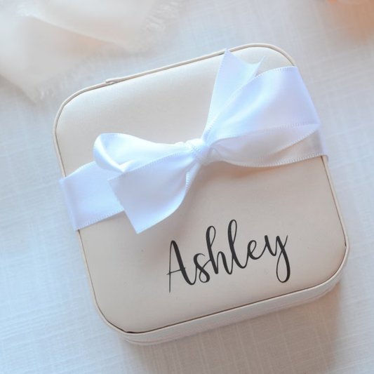 A cream-colored square jewelry box with a white satin bow and the name 'Ashley' printed in black script, set against a soft, light beige fabric background.