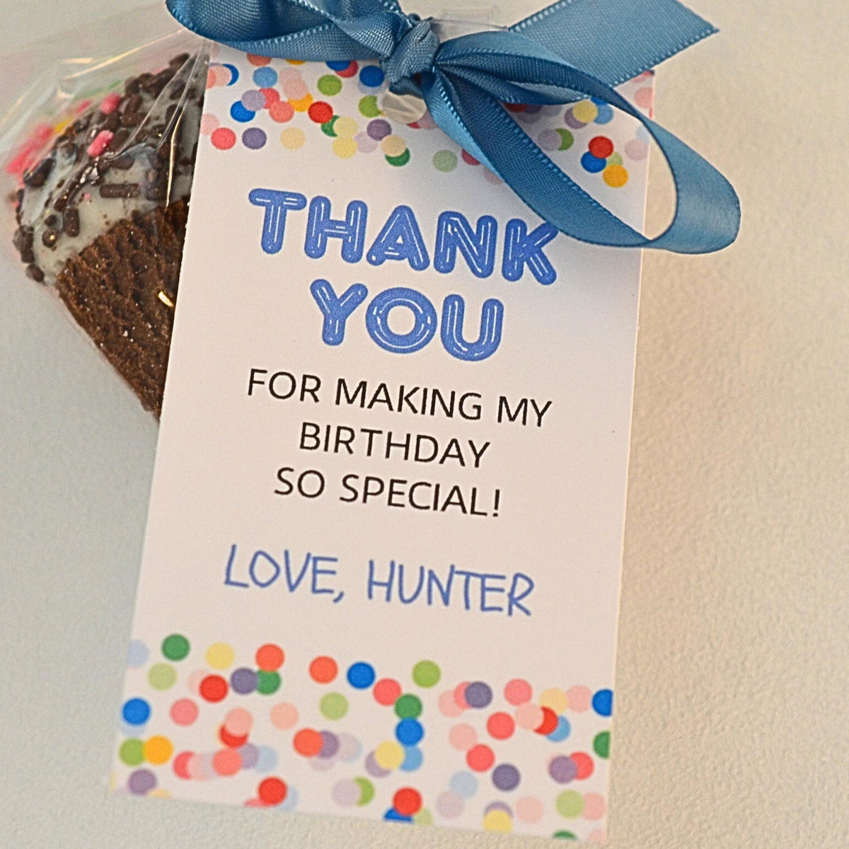 A personalized birthday favor bag containing a chocolate cake, tied with a blue ribbon and featuring a printed white thank you card with colorful confetti borders and blue text.