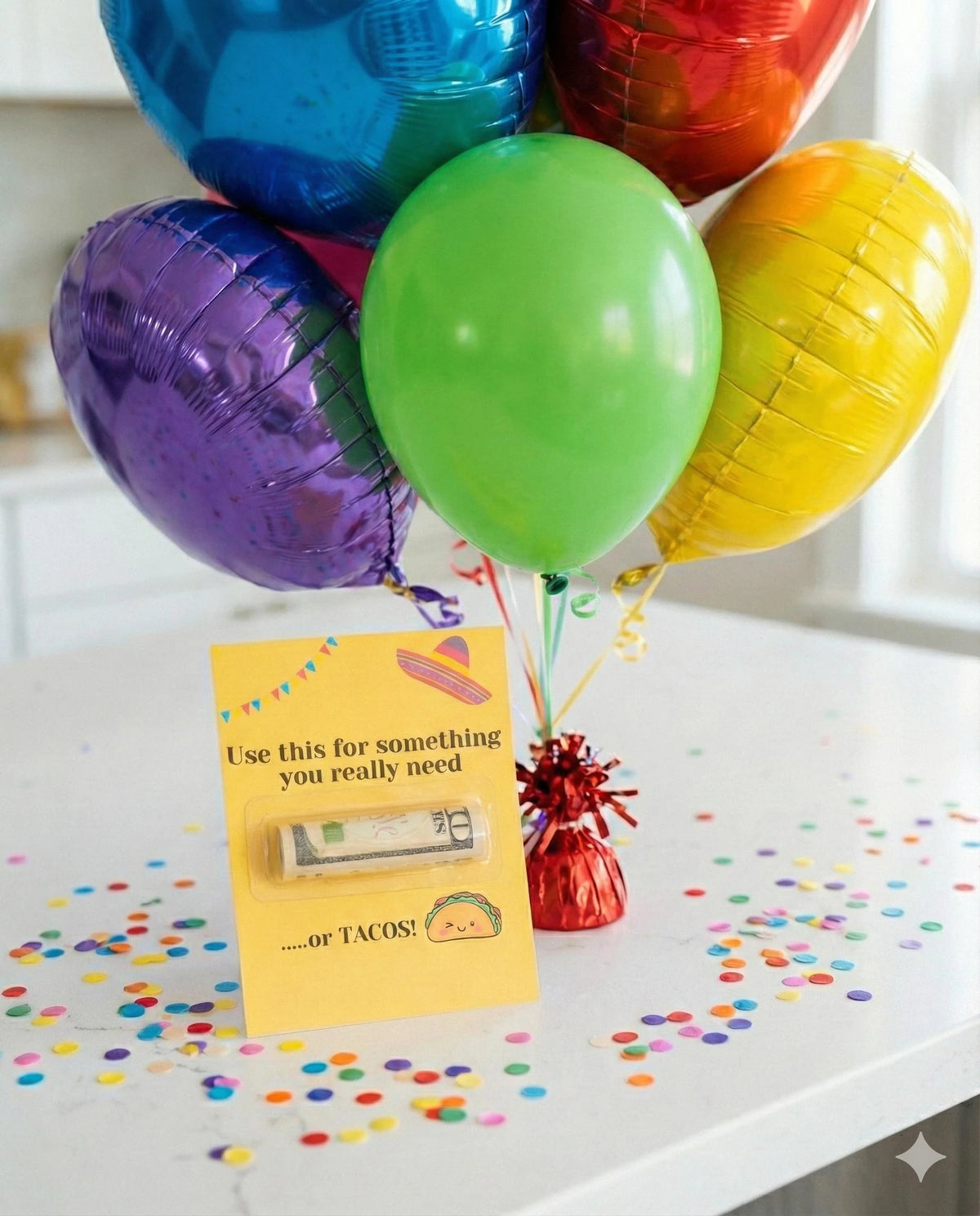 A colorful balloon bouquet with metallic foil balloons in blue, green, purple, yellow, and red, arranged on a white table with confetti. In front of the balloons is a yellow greeting card with a humorous message and a small red tinsel bow.
