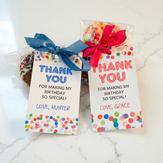 Two packaged birthday treats on a white marble surface, each with a printed thank-you card. The left treat has a blue ribbon and a blue thank-you message, and the right treat has a red ribbon and a pink thank-you message.