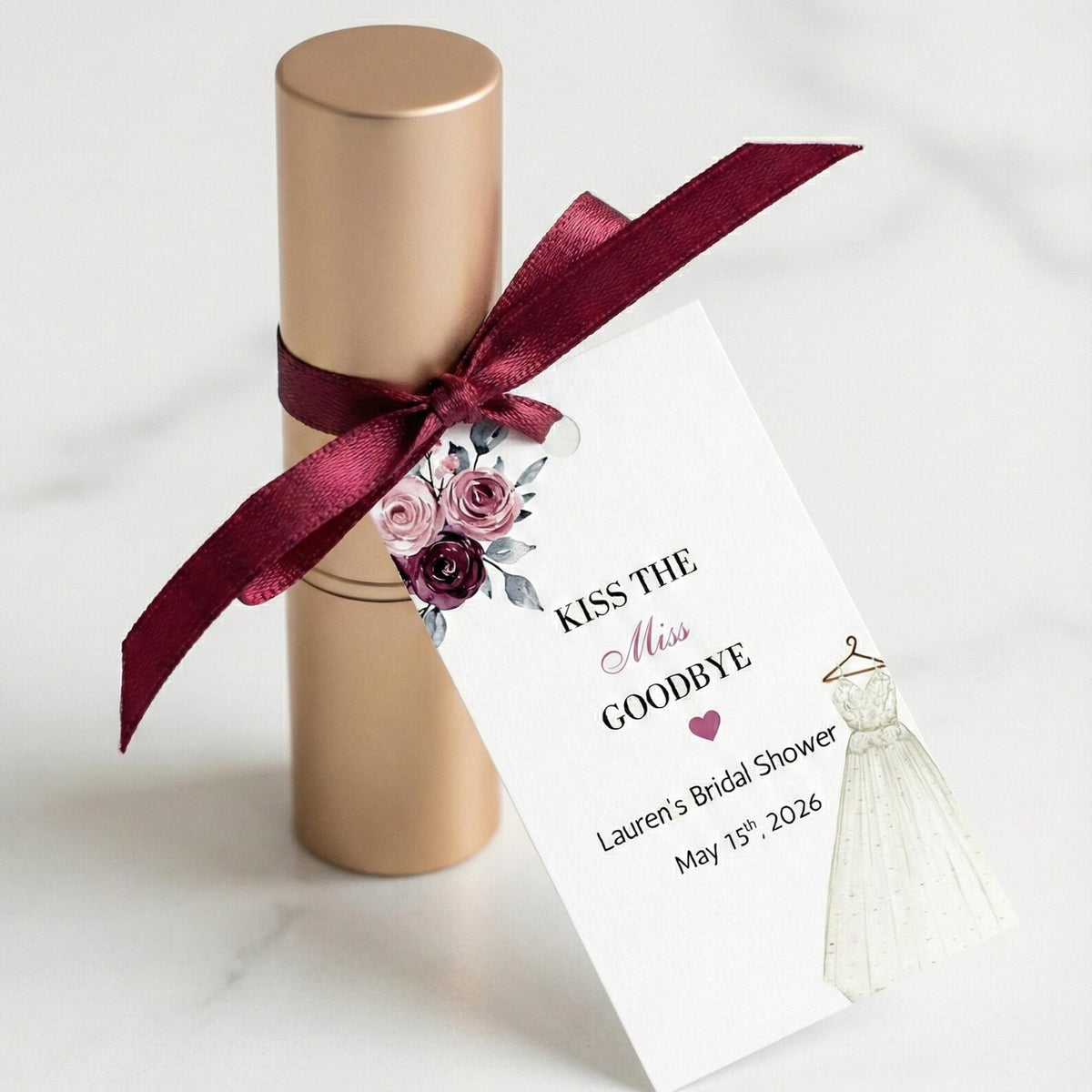 A gold cylindrical gift box tied with a burgundy satin ribbon and a printed wedding-themed tag that reads 'Kiss the Miss Goodbye' with floral artwork and a small bridal gown illustration, photographed on a light marble surface.