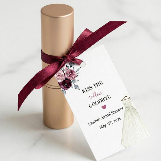 A gold cylindrical gift box tied with a burgundy satin ribbon and a printed wedding-themed tag that reads 'Kiss the Miss Goodbye' with floral artwork and a small bridal gown illustration, photographed on a light marble surface.