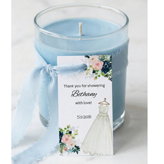 A light blue glass jar candle tied with a pale blue ribbon and a printed shower thank-you tag featuring floral artwork and a white dress illustration, set on a light marble surface.