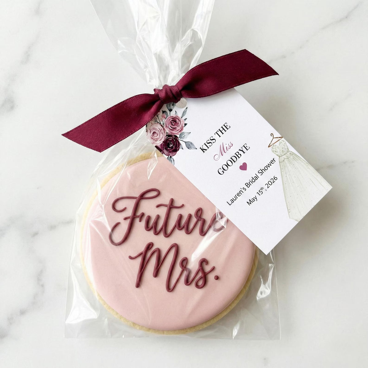 A round sugar cookie with pale pink icing and dark burgundy script reading 'Future Mrs.' is wrapped in clear cellophane with a matching burgundy ribbon and a printed tag that says 'Kiss the Miss Goodbye' and includes a bride illustration.