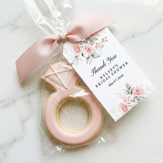 A personalized bridal shower favor featuring a ring-shaped sugar cookie with pale pink icing and a diamond design, wrapped in clear cellophane with a matching pale pink satin ribbon and a floral thank-you card.