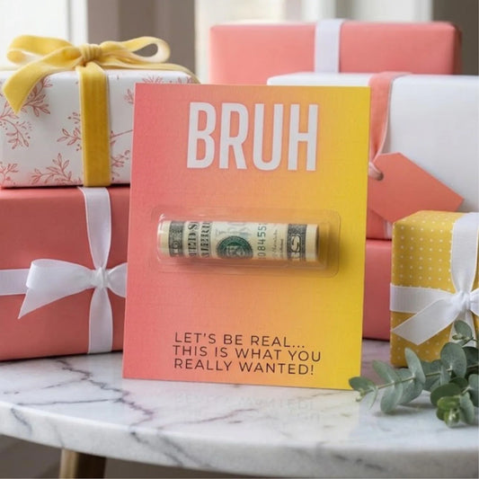 A novelty gift card standing on a marble surface, surrounded by wrapped presents. The card is pink-to-yellow gradient with the word "BRUH" in large white letters and a rolled dollar bill in a clear tube across the center.