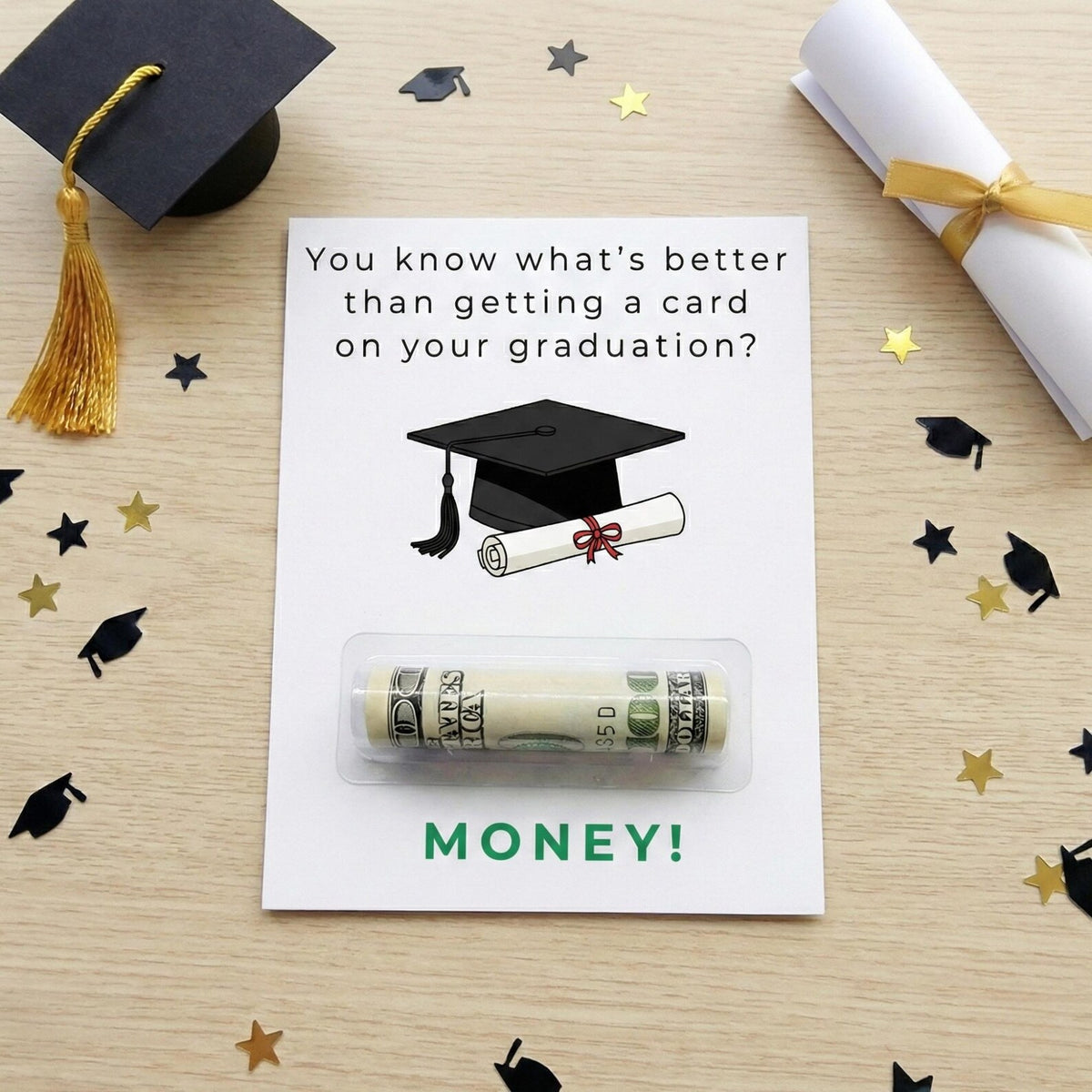 A graduation-themed money card on a light wood tabletop, surrounded by black and gold confetti and styled with a graduation cap, rolled diploma, and gold tassel. The card reads, 'You know what's better than getting a card on your graduation? MONEY!' with an illustration of a graduation cap and rolled diploma above a clear blister pack containing a rolled dollar bill.