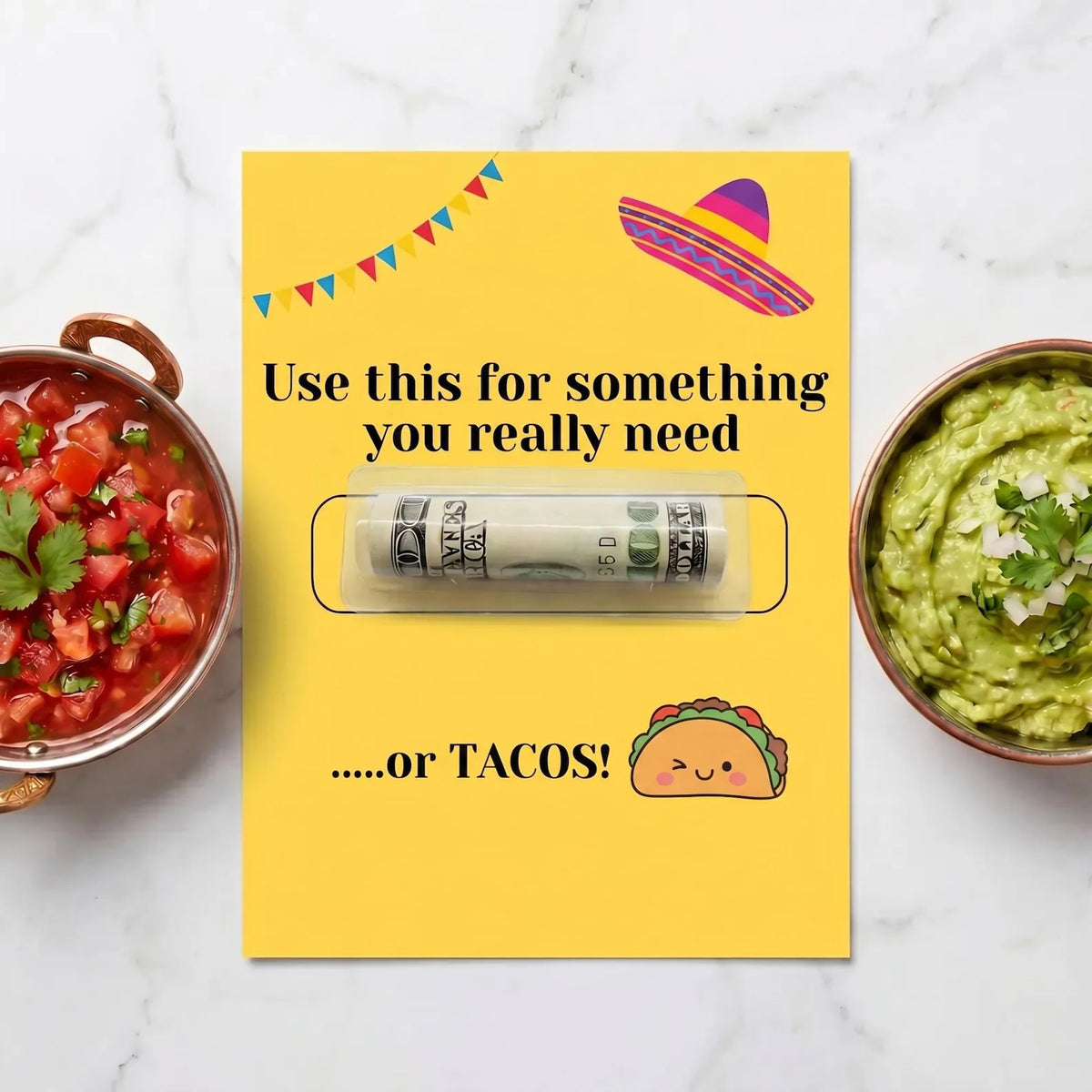 A yellow card or envelope on a white marble surface, featuring a rolled dollar bill tucked in a clear money tube with humorous text. Two bowls of food sit on either side: salsa on the left and guacamole on the right.