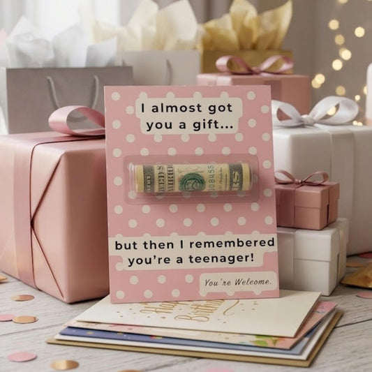 A pink gift card with white polka dots and a humorous message stands in front of wrapped presents. A rolled dollar bill is visible through a clear tube attached to the card.