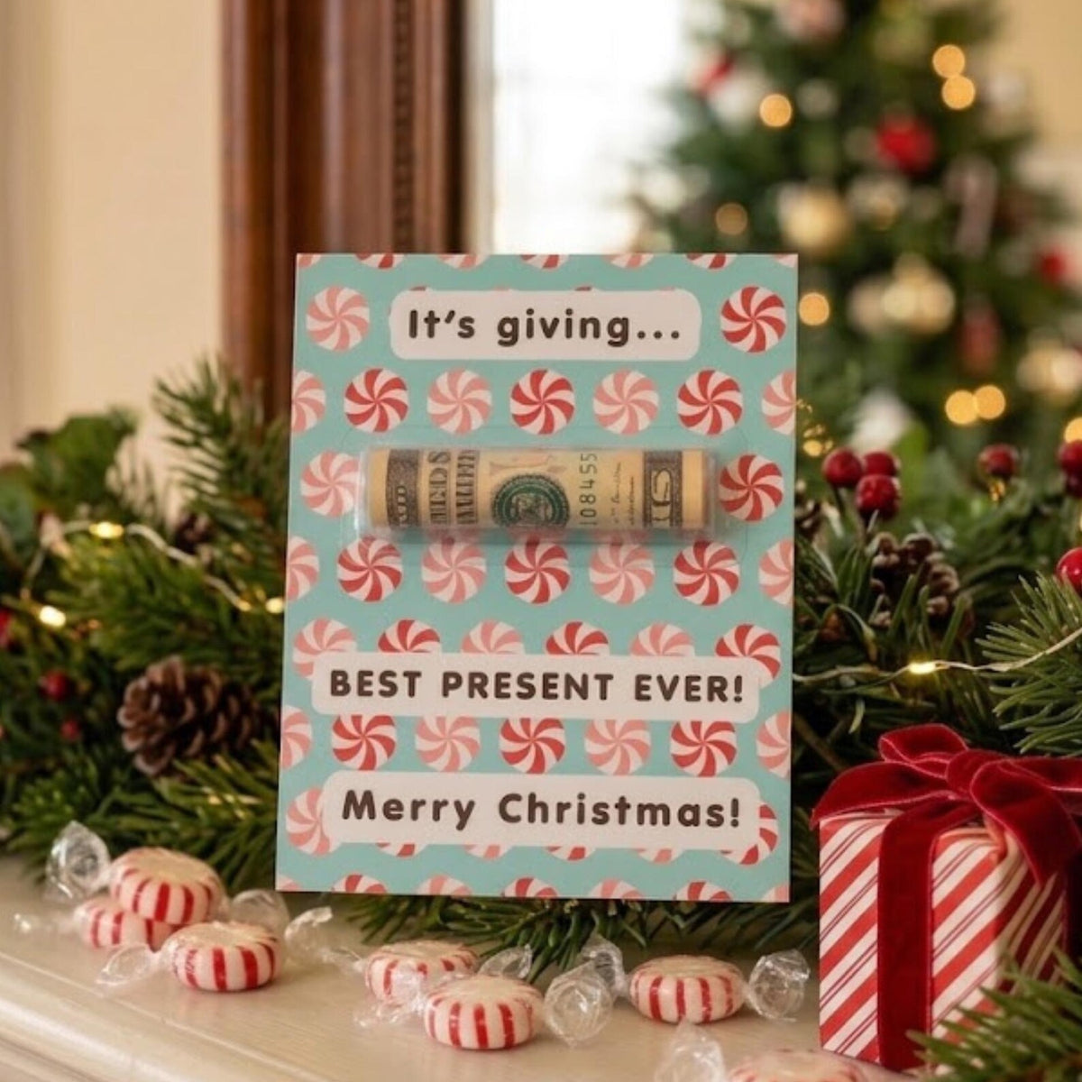 A Christmas-themed gift card with a rolled dollar bill hidden inside, displayed on a mantel with candy, pine branches, and a wrapped gift box. The card reads 'It's giving.