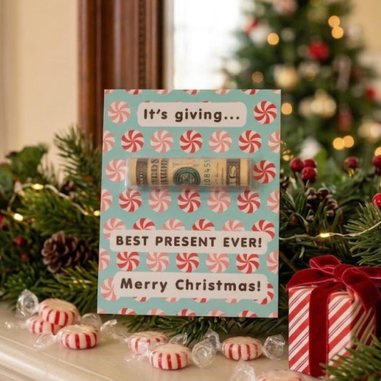 A Christmas-themed gift card with a rolled dollar bill hidden inside, displayed on a mantel with candy, pine branches, and a wrapped gift box. The card reads 'It's giving.